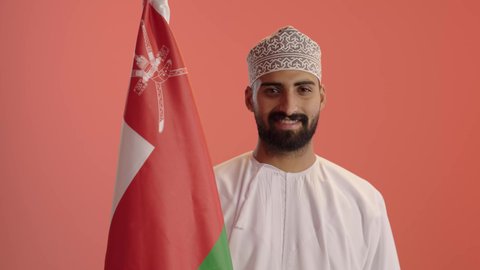 الاعتزاز والفخر بالرموز والراية العمانية، الاحتفال بذكرى اليوم الوطني العماني، المشاركة بالاعياد والمناسبات الوطنية، بورتريه مقرب لشاب عربي خليجي عماني يرتدي الدشداشة والكمه يحمل بيده علم سلطنة عمان، مفهوم حب الوطن والإنتماء إليه، خلفية برتقالي