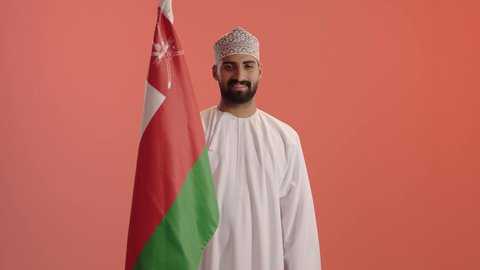 المشاركة بالاعياد والمناسبات الوطنية، الاحتفال بذكرى اليوم الوطني العماني، الاعتزاز والفخر بالرموز والراية العمانية، بورتريه لشاب عربي خليجي عماني يرتدي الدشداشة والكمه يحمل بيده علم سلطنة عمان، مفهوم حب الوطن والإنتماء إليه، خلفية برتقالي