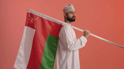 مشاعر الانتماء والفخر بالرموز الوطنية، بورتريه من الجانب لرجل عربي خليجي عماني يرتدي الدشداشة و الكمة يحمل يحمل علم وطنه، الرايات والأعلام الوطنية للدول العربية، النظر الى الكاميرا بايماءات السعادة و السرور، خلفية برتقالي
