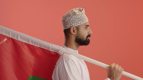 النظر الى الكاميرا بايماءات السعادة و السرور، الرايات والأعلام الوطنية للدول العربية، الفخر والاعتزاز بالرموز والراية العمانية، بورتريه مقرب من الجانب لرجل عربي خليجي عماني يرتدي الدشداشة و الكمة يحمل يحمل علم وطنه، خلفية برتقالي