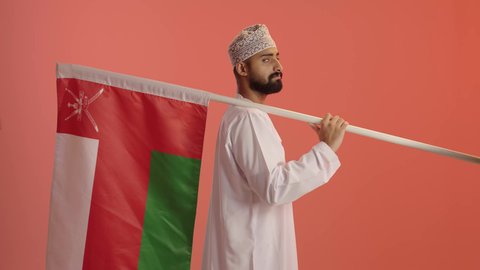 الرايات والأعلام الوطنية للدول العربية، الفخر والاعتزاز بالرموز والراية العمانية، بورتريه من الجانب لرجل عربي خليجي عماني يرتدي الدشداشة و الكمة يحمل يحمل علم وطنه، النظر الى الكاميرا بايماءات السعادة و السرور، خلفية برتقالي
