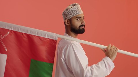 الفخر والاعتزاز بالرموز والراية العمانية، بورتريه من الجانب لرجل عربي خليجي عماني يرتدي الدشداشة و الكمة يحمل يحمل علم وطنه، الرايات والأعلام الوطنية للدول العربية، النظر الى الكاميرا بايماءات السعادة و السرور، خلفية برتقالي
