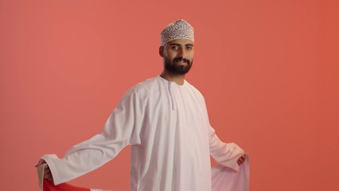 Pride and honor in belonging to the Sultanate of Oman, expressing love and loyalty to the Omani flag, a portrait of an Arab Gulf Omani man wearing a dishdasha and kumma, wrapping the Omani flag around his shoulders and looking at the camera with gestures of happiness and joy, orange background.