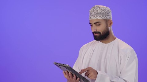 Browsing social media, using modern technology and techniques, following enjoyable movies and series, chatting with family and friends, an Arab Gulf Omani man wearing a dishdasha and kumma holding a tablet in his hand, gestures of engagement and focus.