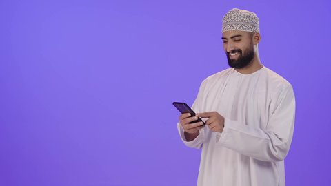الدردشة مع الأهل والأصدقاء، استخدام جهاز تقني حديث، بورتريه لشاب عربي خليجي عماني يرتدي الدشداشة و الكمة يمسك بيده الهاتف المحمول، دمج التقنية والتكنولوجيا بالحياة اليومية، تصفح مواقع التواصل الاجتماعي، ايماءات السعادة و السرور، خلفية بنفسجية
