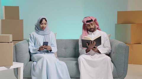 قضاء أوقات ممتعة في قراءة الكتب والروايات، صناديق وكراتين في الخلفية، مفهوم المناقشة والحوار بين الأزواج، بورتريه لرجل عربي خليجي سعودي مثقف يرتدي الثوب و المشاغ  يقوم بتصفح كتاب بجانبه زوجته تحمل الهاتف المحمول، مفهوم التعلم الذاتي والثقافة، خلفية زرقاء