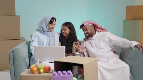 ايماءات السعادة و السرور، مشاهدة الافلام والبرامج الترفيهيه، القيام بالانشطة المسلية في المنزل، قضاء وقت ممتع مع الأبناء، بورتريه لرجل عربي خليجي سعودي يرتدي الثوب التقليدي والشماغ يجلس على الكنبة بجانب ابنته وزوجته يقومون باجراء مكالمة فيديو عبر الحاسوب المحمول، خلفية زرقاء