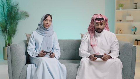 قضاء وقت ممتع بلعب ألعاب الفيديو بإيماءات تدل على السعادة، المتعة و التسلية بألعاب البلايستيشن الترفيهية، مفهوم الأسرة السعيدة، بورتريه لزوجان عربيان خليجيان سعوديان يجلسان في غرفة المعيشة يمسكان عصا التحكم ويلعبان بألعاب الفيديو بتنافس وحذر، خلفية زرقاء