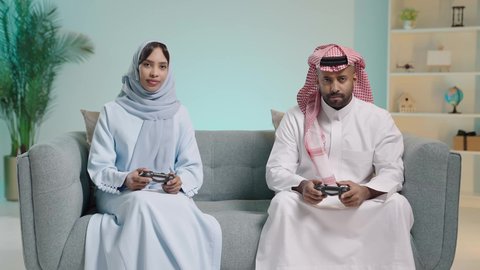 المتعة و التسلية بألعاب البلايستيشن الترفيهية، مفهوم الأسرة السعيدة، قضاء وقت ممتع بلعب ألعاب الفيديو بإيماءات تدل على السعادة، بورتريه لزوجان عربيان خليجيان سعوديان يجلسان في غرفة المعيشة يمسكان عصا التحكم ويلعبان بألعاب الفيديو بتنافس وحذر، خلفية زرقاء
