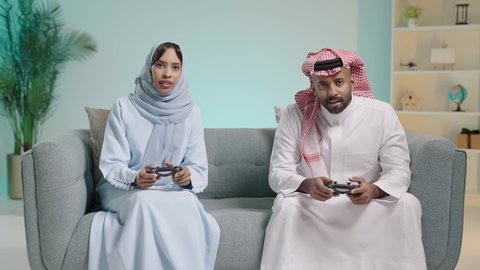 The concept of a happy family, enjoyment and entertainment with recreational PlayStation games, spending quality time playing video games with gestures indicating happiness, a portrait of an Arab Gulf couple from Saudi Arabia sitting in the living room holding a game controller and playing video games competitively and cautiously, with a blue background.