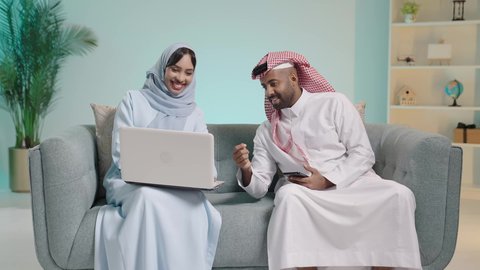 التفاهم وتبادل النقاش بين الأزواج، صناديق وكراتين فى الخلفية، استخدام الأجهزة التقنية الحديثة, بورتريه لرجل عربي خليجي سعودي يرتدي الثوب ويجلس على آريكة وبجابنبه زوجته تعمل على الحاسب المحمول، تصفح برامج التواصل الاجتماعي، خلفية زرقاء