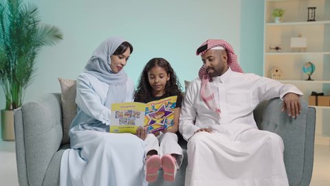 The concept of education and scientific research, explaining and interpreting the events of the story in an interesting and enjoyable way, developing children's skills, the hobby of reading and exploration, spending free time learning and gaining knowledge, a portrait of a Gulf Arab Saudi girl sitting on a couch with her parents reading a book, blue background.