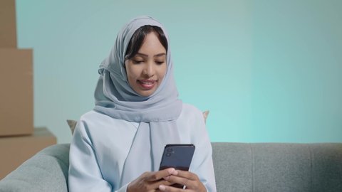 Gestures of integration and focus, online shopping and online orders, chatting and conversations and social communication, conducting a business meeting online, a close-up portrait of a Saudi Arabian Gulf woman with a dark complexion sitting on a sofa and using a mobile phone, blue background.