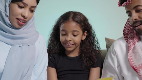 شرح وتفسير أحداث القصة بأسلوب شيق وممتع، تنمية مهرات الطفل، هواية المطالعة والاستكشاف، قضاء وقت الفراغ بالتعلم والثقافة، مفهوم التثقيف والبحث العلمي، بورتريه مقرب  لبنت عربيه خليجيه سعودية تجلس على الاريكه برفقه والديها تقوم بقراءة كتاب، خلفية زرقاء