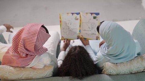 تنمية مهرات الطفل، هواية المطالعة والاستكشاف، قضاء وقت الفراغ بالتعلم والثقافة، مفهوم التثقيف والبحث العلمي، بورتريه مقرب من الخلف لبنت عربيه خليجيه سعودية تجلس على الاريكه برفقه والديها تقوم بقراءة كتاب ، شرح وتفسير أحداث القصة بأسلوب شيق وممتع، خلفية زرقاء