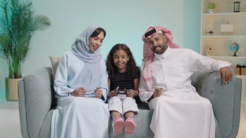 قضاء وقت ممتع بلعب ألعاب الفيديو، المتعة و التسلية بألعاب البلايستيشن الترفيهية، مفهوم الأسرة السعيدة، بورتريه لبنت عربيه خليجيه سعودية ترتدي الزي الكاجوال تجلس في غرفة المعيشة برفقه والديها تمسك عصا التحكم تلعب بألعاب الفيديو بتنافس وحذر، ايماءات الفوز و الانتصار ، خلفية زرقاء