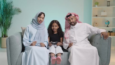 المتعة و التسلية بألعاب البلايستيشن الترفيهية، مفهوم الأسرة السعيدة، قضاء وقت ممتع بلعب ألعاب الفيديو، بورتريه لبنت عربيه خليجيه سعودية ترتدي الزي الكاجوال تجلس في غرفة المعيشة برفقه والديها تمسك عصا التحكم تلعب بألعاب الفيديو بتنافس وحذر،ايماءات الفوز و الانتصار ، خلفية زرقاء