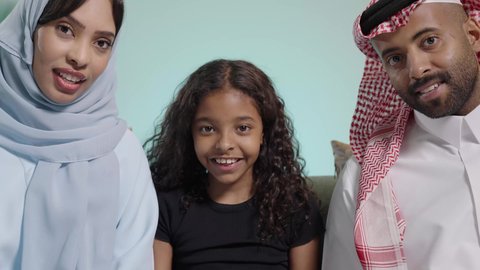 مفهوم الأسرة السعيدة، المتعة و التسلية بألعاب البلايستيشن الترفيهية، قضاء وقت ممتع بلعب ألعاب الفيديو، بورتريه مقرب لبنت عربيه خليجيه سعودية ترتدي الزي الكاجوال تجلس في غرفة المعيشة برفقه والديها تمسك عصا التحكم تلعب بألعاب الفيديو بتنافس وحذر،ايماءات الفوز و الانتصار ، خلفية زرقاء