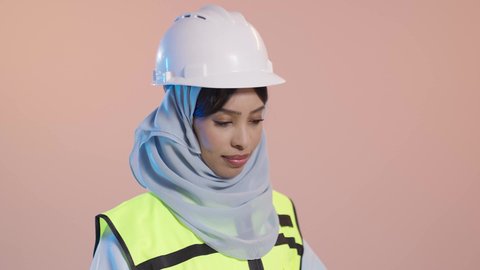 Verification of the implementation of the construction project, the concept of engineering and architectural construction, a profession and job for Saudi women, a close portrait of a Saudi Gulf Arab female engineer wearing a hijab, an abaya, a jacket, and a safety helmet holding the engineering plan in her hand, with a pink background.