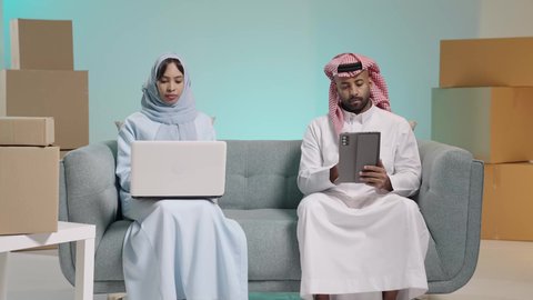 تصفح برامج التواصل الاجتماعي، تطبيقات الأجهزة الحاسوبية، صناديق وكراتين في الخلفية، استخدام الأجهزة التقنية الحديثة, بورتريه لرجل عربي خليجي سعودي يرتدي الثوب يجلس على آريكة يحمل بيده جهاز التابلت بجانب زوجته تعمل على الحاسب المحمول، خلفية زرقاء