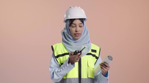 المتابعة والإشراف على المشاريع الهندسية، بورتريه مقرب لمهندسة عربية خليجية سعودية سمراء  ترتدي الحجاب  وسترة الحماية و الخوذه تحمل جهاز اتصال لاسلكي و المخطط الورقي ، مهن ووظائف شبابية، إعطاء أوامر خاصة بالعمل، خلفية وردي