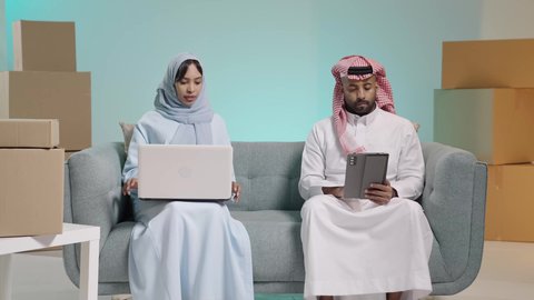 تطبيقات الأجهزة الحاسوبية، استخدام الأجهزة التقنية الحديثة، صناديق وكراتين في الخلفية،  التحاور والنقاش في الرأي مع الأزواج, بورتريه لرجل عربي خليجي سعودي يرتدي الثوب يجلس على آريكة يحمل بيده جهاز التابلت بجانب زوجته تعمل على الحاسب المحمول، تصفح برامج التواصل الاجتماعي، خلفية زرقاء