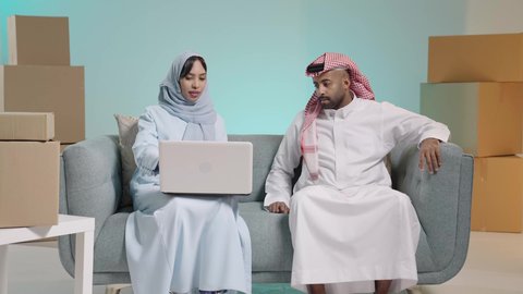 استخدام الأجهزة التقنية الحديثة، التحاور والنقاش في الرأي مع الأزواج, صناديق وكراتين في الخلفية، بورتريه لرجل عربي خليجي سعودي يرتدي الثوب يجلس على آريكة بجانب زوجته تعمل على الحاسب المحمول، تصفح برامج التواصل الاجتماعي، تطبيقات الأجهزة الحاسوبية، خلفية زرقاء