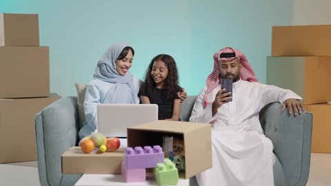 القيام بالانشطة المسلية في المنزل، مشاهدة الافلام والبرامج الترفيهيه، قضاء وقت ممتع مع الأبناء، بورتريه لام عربيه خليجيه سعودية ترتدي العباءه تجلس على الكنبة بجانب ابنتها يقومون باجراء مكالمة فيديو عبر الحاسوب المحمول، ، صناديق وكراتين في الخلفية، رجل عربي خليجي سعودي يقوم بتصفح الهاتف المحمول ، خلفية زرقاء