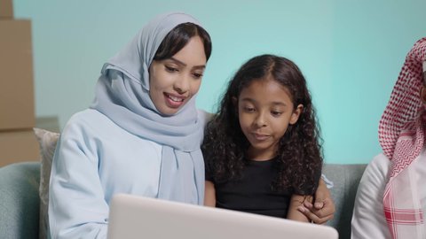 Watching movies and entertainment programs, doing fun activities at home, spending quality time with children, a portrait of an Arab Gulf Saudi man wearing traditional dress and a headscarf sitting on the sofa next to his daughter and wife making a video call via a laptop, blue background.