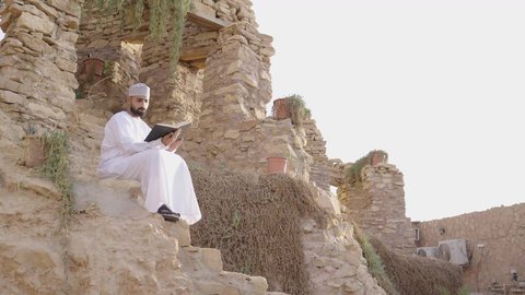 مراجعة البحوث العلمية، قضاء وقت ممتع في قراءة الكتب والروايات، تنمية الذخيرة اللغوية والعلمية، شاب عربي خليجي عماني مثقف يرتدي الدشداشة والكمة يجلس في إحدى الحدائق العامة يقوم بقراءة كتاب، مفهوم التعلم الذاتي والاطلاع والثقافة، الشغف في ممارسة هواية المطالعة