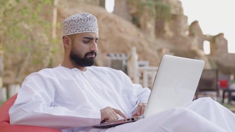إنجاز مهام العمل عن بعد عبر اللابتوب، شاب عربي خليجي عماني مثقف يرتدي الدشداشة والكمة يجلس في إحدى الحدائق العامة يقوم باستخدام الحاسوب المحمول، قضاء وقت ممتع في الهواء الطلق، تصفح الانترنت ومواقع السوشال ميديا