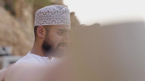 قضاء وقت ممتع في الهواء الطلق، تصفح الانترنت ومواقع السوشال ميديا، إنجاز مهام العمل عن بعد عبر اللابتوب، تصوير مقرب لشاب عربي خليجي عماني مثقف يرتدي الدشداشة والكمة يجلس في إحدى الحدائق العامة يقوم باستخدام الحاسوب المحمول