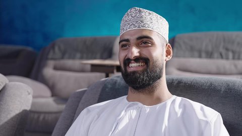 الشغف في ممارسة الهوايات، قضاء وقت ممتع في خارج المنزل، مشاهدة فلم سينمائي، تصوير مقربة  لرجل عربي خليجي عماني يرتدي الثوب التقليدي يجلس على الكنبة ينظر إلى مكان ما بايماءات السعادة والسرور، انشطة ترفيهية خارجية، تعابير الاندماج والتركيز، متابعة برامج التلفاز