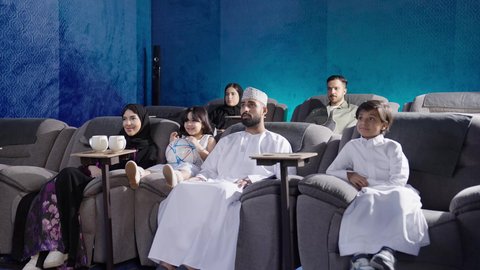 مقاعد سينما مريحة وعصرية، مشاهدة فيلم سينمائي داخل قاعة السينما، قضاء أوقات ممتعة، الاستمتاع بمشاهدة المباريات في عطلة نهاية الأسبوع، عائلة عربية خليجية عمانية أم ترتدي الحجاب والعباءة تجلس في السينما مع زوجها وطفلتها، نزهات الأصدقاء لقضاء أوقات ممتعة
