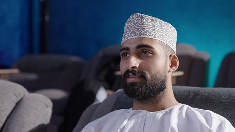 انشطة ترفيهية خارجية،الشغف في ممارسة الهوايات، قضاء وقت ممتع في خارج المنزل، مشاهدة فلم سينمائي، تصوير مقربة  لرجل عربي خليجي عماني يرتدي الثوب التقليدي يجلس على الكنبة ينظر إلى مكان ما بايماءات السعادة والسرور، تعابير الاندماج والتركيز، متابعة برامج التلفاز