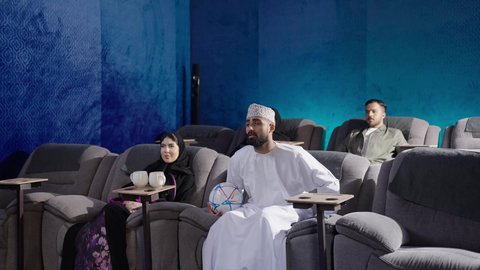 نزهات الأصدقاء معا لقضاء أوقات ممتعة، الاستمتاع بمشاهدة المباريات في عطلة نهاية الأسبوع،بناء علاقات اجتماعية ناجحة، إمرأة عربية خليجية عمانية ترتدي الحجاب والعباءة تجلس في السينما ترفع يديها بإيماءات البهجة والسرور، مشاهدة فيلم سينمائي، قضاء أوقات ممتعة ومسلية