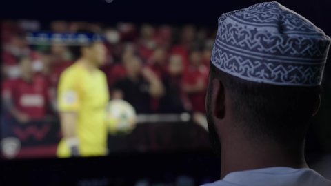 مشاهدة المباراة والحماس , الشغف بالانشطة الترفيهية، ايماءات الفوز والانتصار ، المباريات والألعاب الرياضية، رجل عربي خليجي عماني يرتدي الزى التقليدى يجلس على أريكة مريحة ، المشاهدة بتعابير التركيز والاندماج ، متابعة مباراة كرة القدم .