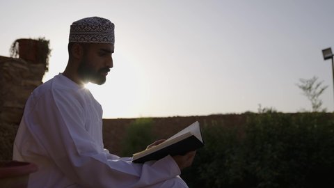 تنمية الذخيرة اللغوية والعلمية، شاب عربي خليجي عماني مثقف يرتدي الدشداشة والكمة يجلس في إحدى الحدائق العامة يقوم بقراءة كتاب، مفهوم التعلم الذاتي والاطلاع والثقافة، قضاء أوقات ممتعة في قراءة الكتب والقصص والروايات، الشغف في ممارسة هواية المطالعة