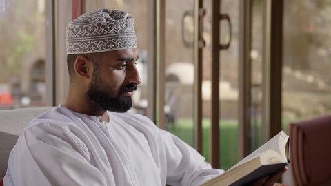دعم وتنمية الذخيرة اللغوية والعلمية، تصوير مقرب لشاب عربي خليجي عماني مثقف يقوم بتصفح كتاب، مفهوم التعلم الذاتي والاطلاع والثقافة، قضاء أوقات ممتعة في قراءة الكتب والقصص والروايات، جلسة في إحدى الشاليهات الخارجية، الشغف في ممارسة هواية المطالعة