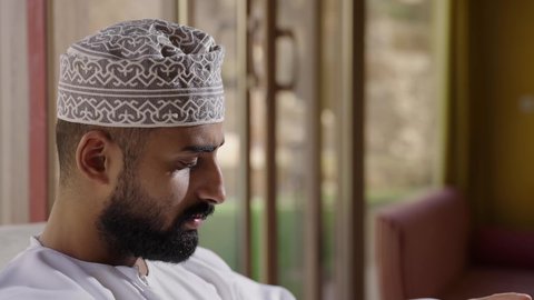 مفهوم التعلم الذاتي والاطلاع والثقافة، قضاء أوقات ممتعة في قراءة الكتب والقصص والروايات، جلسة في إحدى الشاليهات الخارجية، الشغف في ممارسة هواية المطالعة، دعم وتنمية الذخيرة اللغوية والعلمية، تصوير مقرب لشاب عربي خليجي عماني مثقف يرتدي الدشداشة والكمةيقوم بتصفح كتاب