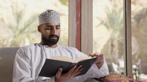 جلسة في إحدى الشاليهات الخارجية، الشغف في ممارسة هواية المطالعة، دعم وتنمية الذخيرة اللغوية والعلمية، قضاء أوقات ممتعة في قراءة الكتب والقصص والروايات، تصوير مقرب لشاب عربي خليجي عماني مثقف يقوم بتصفح كتاب، مفهوم التعلم الذاتي والاطلاع والثقافة