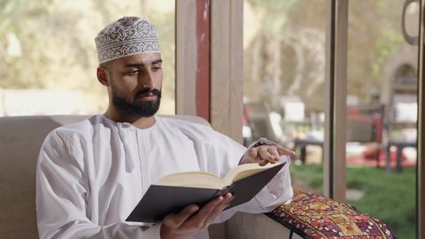 الشغف في ممارسة هواية المطالعة، دعم وتنمية الذخيرة اللغوية والعلمية، قضاء أوقات ممتعة في قراءة الكتب والقصص والروايات، تصوير مقرب لشاب عربي خليجي عماني مثقف يرتدي الدشداشة والكمة يقوم بتصفح كتاب، مفهوم التعلم الذاتي والاطلاع والثقافة