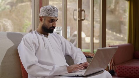 ايماءات الاندماج والتركيز، جهاز تقني حديث ومتطور، إنجاز مهام العمل عن بعد عبر اللابتوب، شاب عربي خليجي عماني مثقف يرتدي الدشداشة والكمة يجلس في إحدى الشاليهات يقوم باستخدام الحاسوب المحمول، تصفح الانترنت ومواقع السوشال ميديا