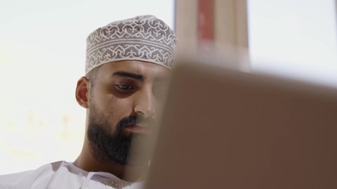 تصفح الانترنت ومواقع السوشال ميديا، ايماءات الاندماج والتركيز، جهاز تقني حديث ومتطور، إنجاز مهام العمل عن بعد عبر اللابتوب، تصوير مقرب لشاب عربي خليجي عماني مثقف يرتدي الدشداشة والكمة يجلس في إحدى الشاليهات يقوم باستخدام الحاسوب المحمول