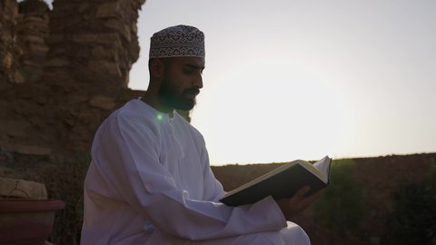 تنمية الذخيرة اللغوية والعلمية، شاب عربي خليجي عماني مثقف يرتدي الدشداشة والكمة يجلس في إحدى الحدائق العامة يقوم بقراءة كتاب، مفهوم التعلم الذاتي والاطلاع والثقافة، قضاء أوقات ممتعة في قراءة الكتب والقصص والروايات، الشغف في ممارسة هواية المطالعة