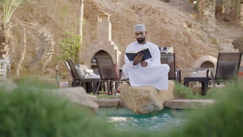 قضاء أوقات ممتعة في قراءة الكتب والقصص والروايات، تنمية الذخيرة اللغوية والعلمية، شاب عربي خليجي عماني مثقف يرتدي الدشداشة والكمة يجلس في إحدى الحدائق العامة يقوم بقراءة كتاب، مفهوم التعلم الذاتي والاطلاع والثقافة، الشغف في ممارسة هواية المطالعة