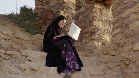 إنجاز مهام العمل عن بعد، استخدام التقنيات الحديثة، اجراء اجتماع عمل عبر الانترنت، دردشة و تواصل اجتماعي ، التسوق الالكتروني وطلبات الاونلاين ، إمرأة عربية عمانية خليجية ترتدي العباءة والحجاب تستخدم الحاسب المحمول وبجانبها زوجها يجلسان في احدى الحدائق العامة