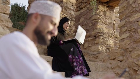 Using modern technologies, conducting a business meeting online, chatting and social communication, completing work tasks remotely, online shopping and orders, an Arab Omani Gulf man wearing traditional attire is using a mobile phone, sitting next to his wife in one of the public parks.