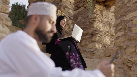 اجراء اجتماع عمل عبر الانترنت، دردشة ومحادثات و تواصل اجتماعي ، إنجاز مهام العمل عن بعد، التسوق الالكتروني وطلبات الاونلاين ، رجل عربي عماني خليجي يرتدي الثوب التقليدي يستخدم الهاتف المحمول وبجانبه زوجته يجلسان في احدى الحدائق العامة، استخدام التقنيات الحديثة