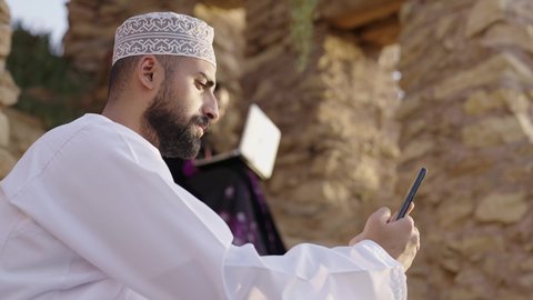 دردشة ومحادثات و تواصل اجتماعي ، اجراء اجتماع عمل عبر الانترنت، إنجاز مهام العمل عن بعد، التسوق الالكتروني وطلبات الاونلاين ، رجل عربي عماني خليجي يرتدي الثوب التقليدي يستخدم الهاتف المحمول وبجانبه زوجته يجلسان في احدى الحدائق العامة، استخدام التقنيات الحديثة